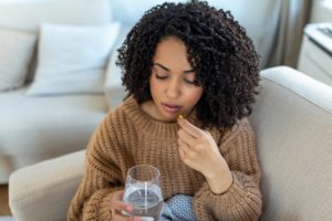 a young black woman sitting on her couch and taking a pill with a glass of water