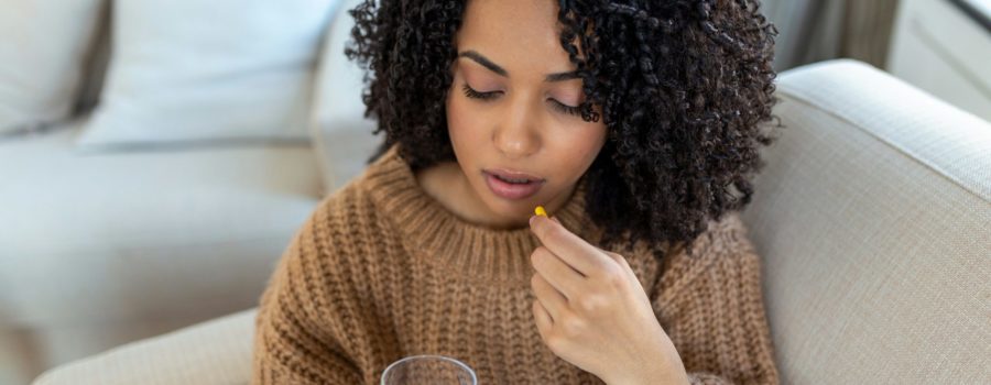 a young black woman sitting on her couch and taking a pill with a glass of water